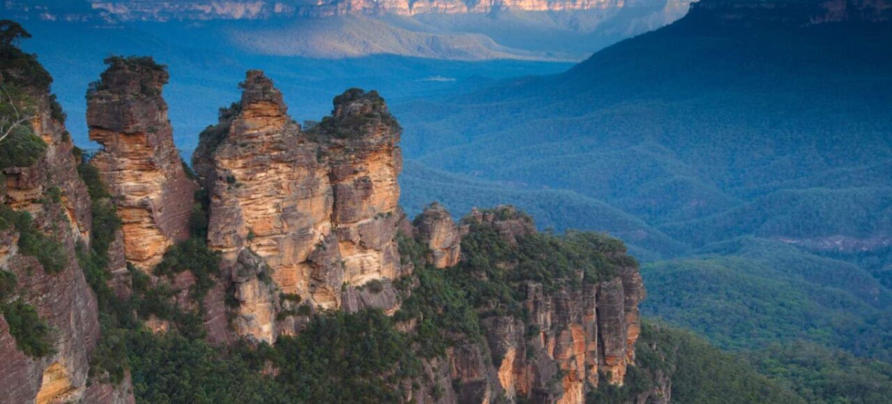 The iconic Three Sisters rock formation in the Blue Mountains, New South Wales, Australia, with vast eucalyptus-covered valleys stretching into the distance under a blue sky.