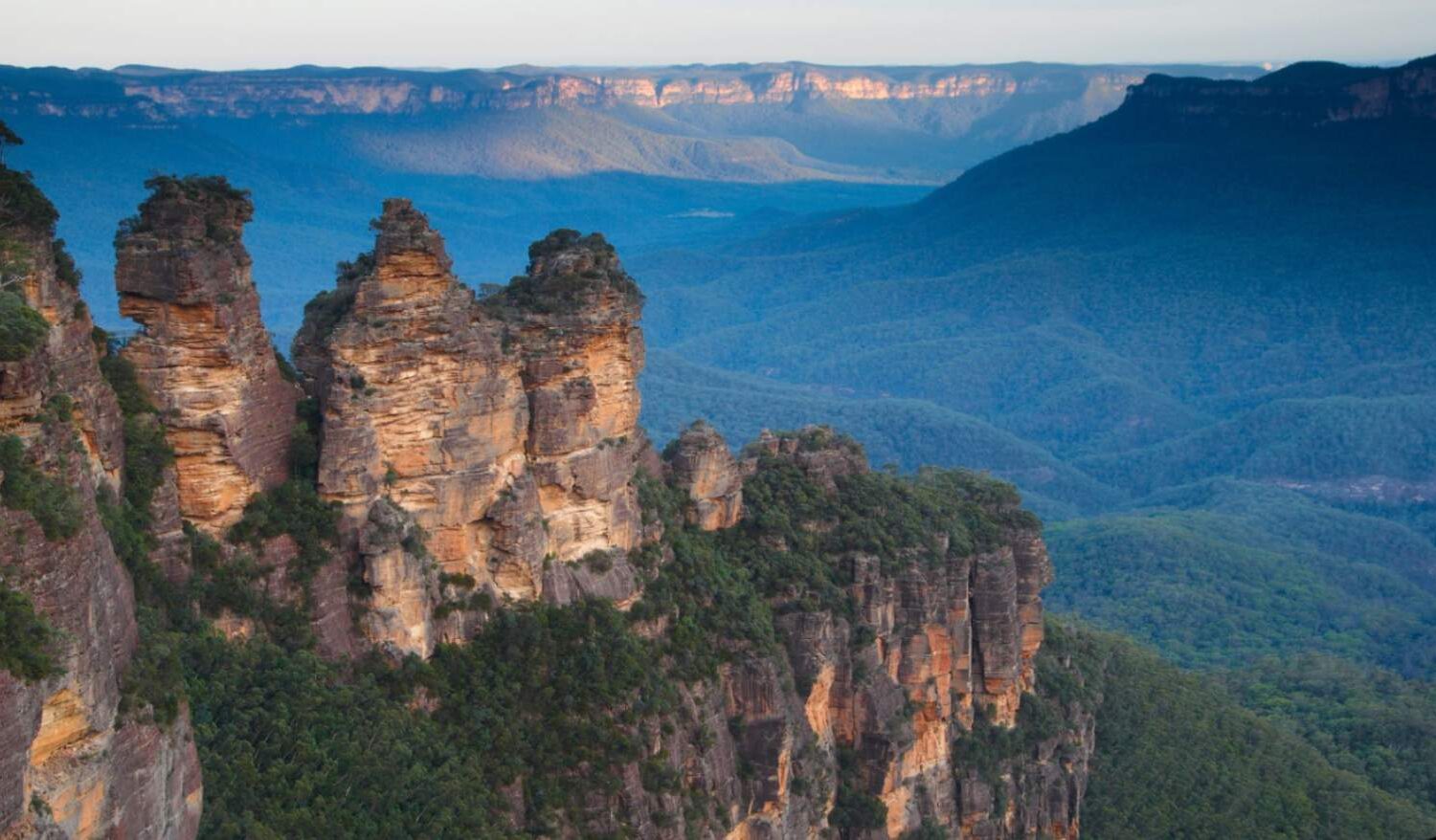 The iconic Three Sisters rock formation in the Blue Mountains, New South Wales, Australia, with vast eucalyptus-covered valleys stretching into the distance under a blue sky.