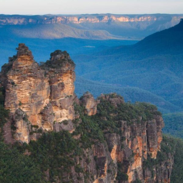 The iconic Three Sisters rock formation in the Blue Mountains, New South Wales, Australia, with vast eucalyptus-covered valleys stretching into the distance under a blue sky.