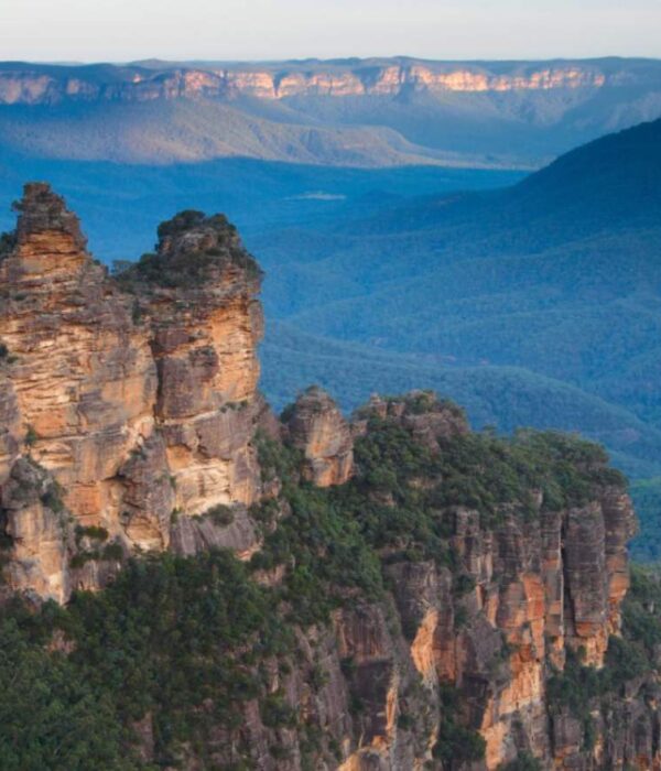 The iconic Three Sisters rock formation in the Blue Mountains, New South Wales, Australia, with vast eucalyptus-covered valleys stretching into the distance under a blue sky.