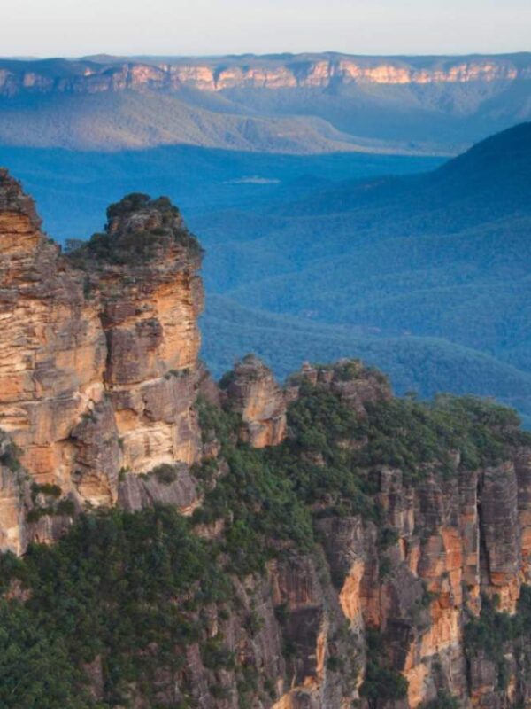The iconic Three Sisters rock formation in the Blue Mountains, New South Wales, Australia, with vast eucalyptus-covered valleys stretching into the distance under a blue sky.