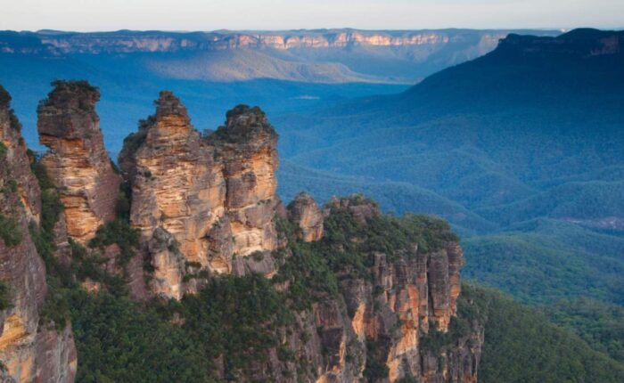 The iconic Three Sisters rock formation in the Blue Mountains, New South Wales, Australia, with vast eucalyptus-covered valleys stretching into the distance under a blue sky.