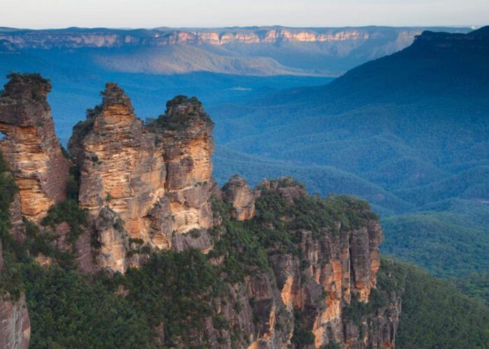 The iconic Three Sisters rock formation in the Blue Mountains, New South Wales, Australia, with vast eucalyptus-covered valleys stretching into the distance under a blue sky.
