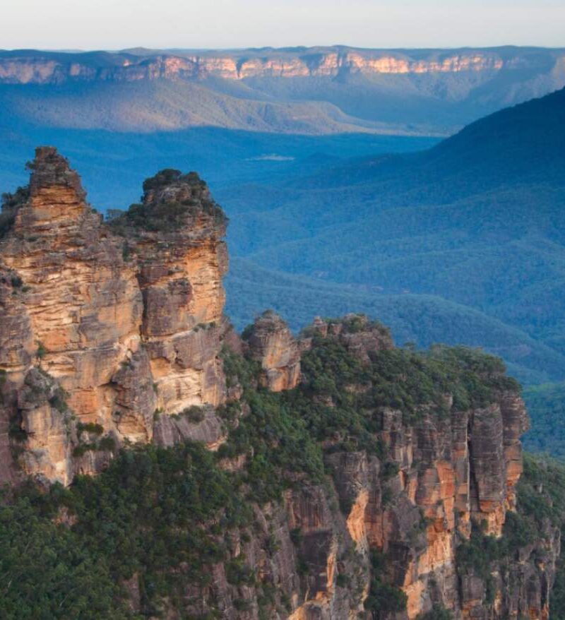 The iconic Three Sisters rock formation in the Blue Mountains, New South Wales, Australia, with vast eucalyptus-covered valleys stretching into the distance under a blue sky.