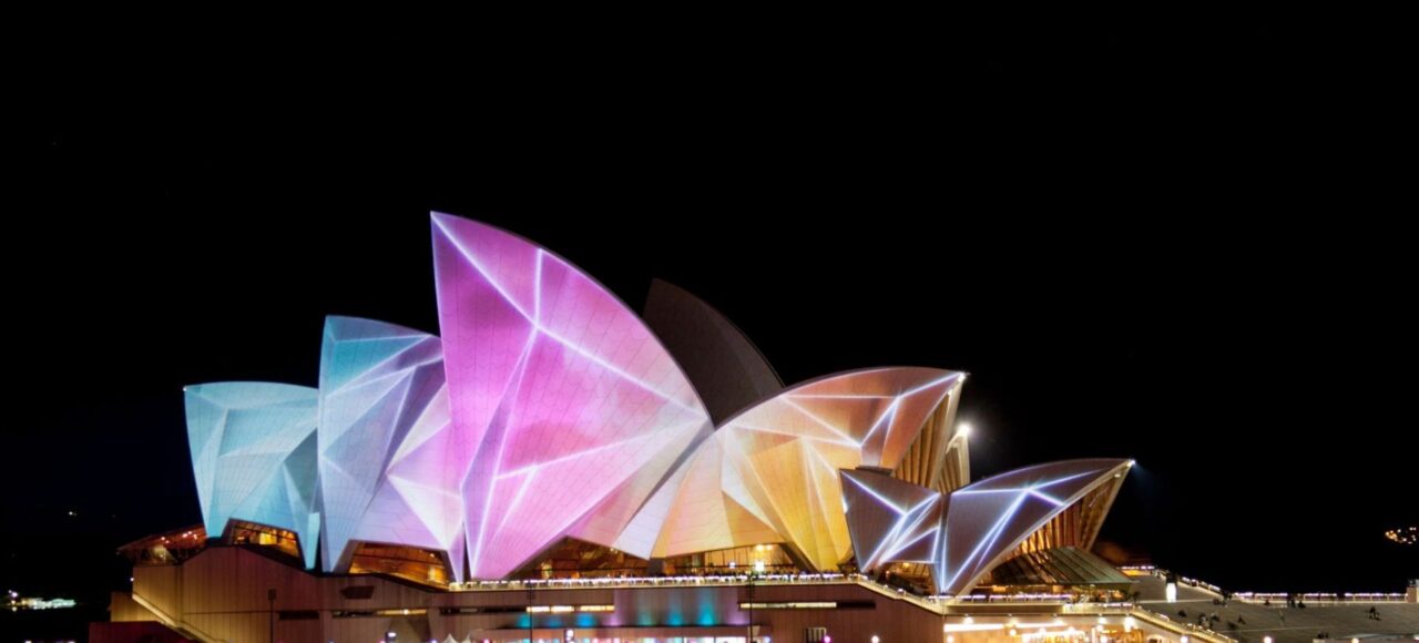 The Sydney Opera House illuminated with vibrant, colorful projections during a night festival, seen from across the water.