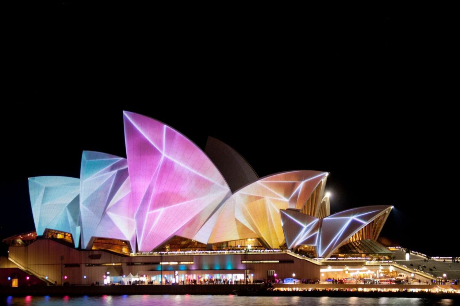 The Sydney Opera House illuminated with vibrant, colorful projections during a night festival, seen from across the water.