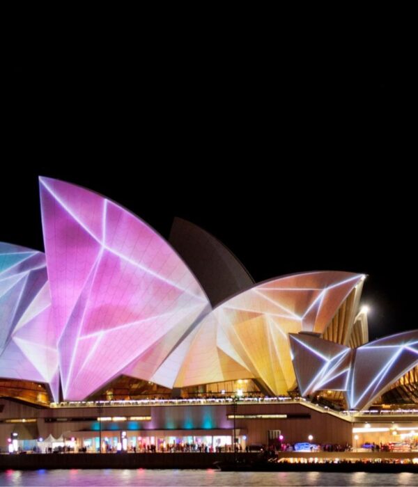 The Sydney Opera House illuminated with vibrant, colorful projections during a night festival, seen from across the water.