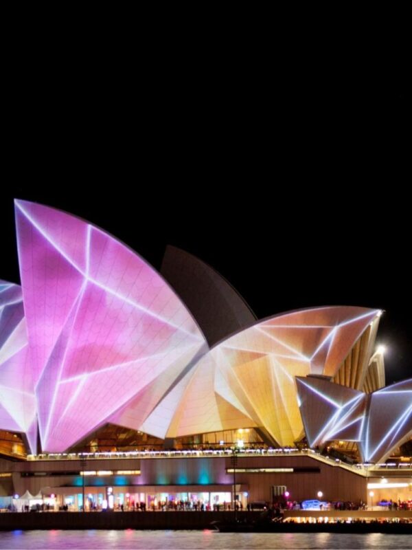 The Sydney Opera House illuminated with vibrant, colorful projections during a night festival, seen from across the water.