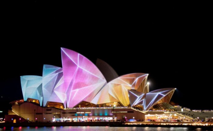 The Sydney Opera House illuminated with vibrant, colorful projections during a night festival, seen from across the water.