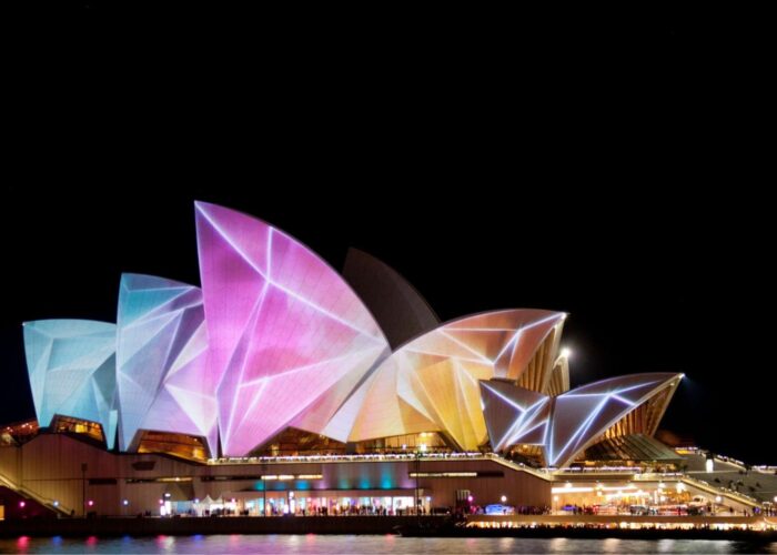 The Sydney Opera House illuminated with vibrant, colorful projections during a night festival, seen from across the water.