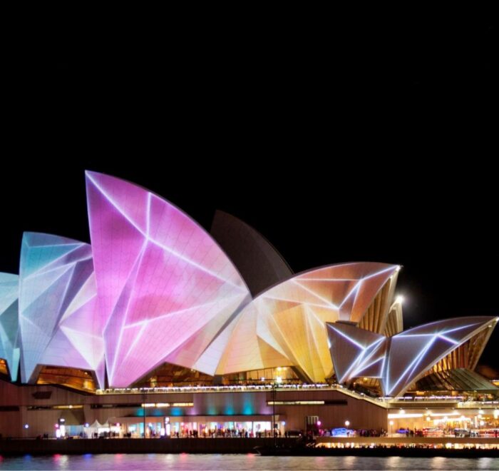 The Sydney Opera House illuminated with vibrant, colorful projections during a night festival, seen from across the water.